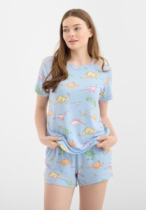 Młoda kobieta ubrana w jasnoniebieskie dopasowane szorty i T-shirt z kolorowym nadrukiem dinozaura w stylu kreskówkowym, ręce w kieszeniach, patrząca bokiem.