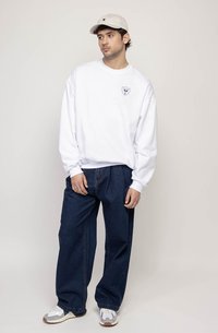 Sweatshirt branco com um pequeno logótipo preto, combinado com jeans de ganga azul-escuro de corte largo e ténis cinzentos e brancos, rematado com um boné bege.