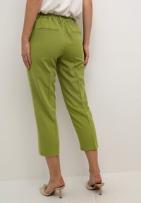Vrouw in cropped olijfgroene broeken met elastische tailleband en beige hoge muiltjes, staand tegen een effen witte achtergrond.
