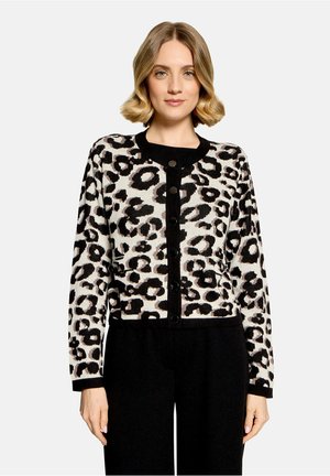 Woman with blonde hair wearing a black and white leopard print buttoned cardigan and black pants, standing against a plain light background.