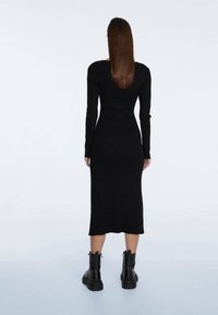 Schwarz, langärmliges, eng anliegendes Strickkleid mit Midi-Länge. Präsentiert einen tiefen Rundhalsausschnitt und gerippte Textur. Getragen mit klobigen schwarzen Stiefeletten.