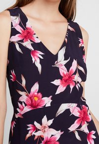 Robe florale avec un fond navy et de grandes fleurs roses et blanches, un décolleté en V et un corsage ajusté. Tissu doux avec une texture lisse.