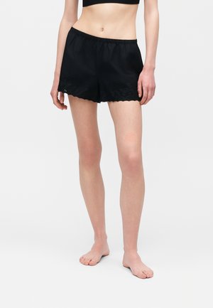 Naine kannab musta elastse vöökohaga lühikesi pükse pitsilise äärisega, seisab paljajalu ühetaustal valgel taustal.