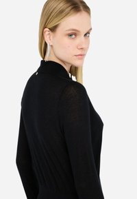 Top nero a maglia con colletto alto, texture a coste e dettaglio plissettato sul retro. Il modello indossa lunghe orecchine scintillanti.