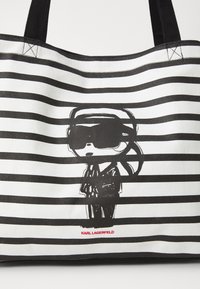 Geantă de pânză cu dungi orizontale negre și albe. Prezintă o figură de desene animate purtând ochelari de soare și costum. Logo-ul roșu "KARL LAGERFELD" în partea de jos.