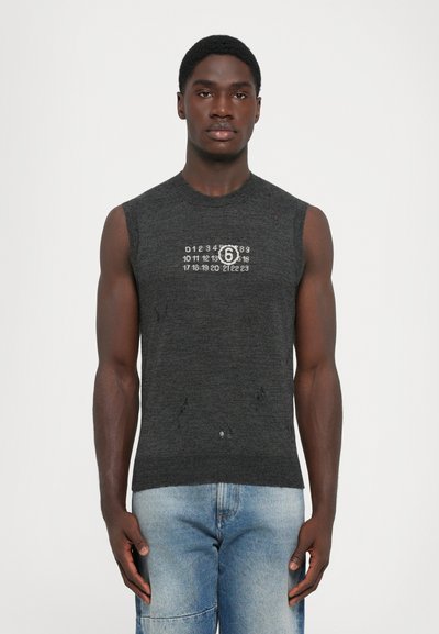 Uomo slanciato che indossa una maglietta senza maniche grigio scuro a maglia con numeri bianchi e buchi strappati, abbinata a jeans azzurri, in piedi su uno sfondo bianco.
