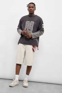 Γκρι φούτερ με το "89" σε άσπρο, γκρι μανίκια. Κρέμα ριγέ σορτς, κόκκινο μαντήλι να φαίνεται. Λευκά αθλητικά παπούτσια με διακριτικό λογότυπο.