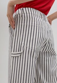 Pruhované nohavice s vysokým pásom v bielej a námornej modri, s chrbtovým vreckom a textúrovanou tkaninou. Ruka na boku opretá o pás.