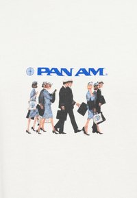 Załoga lotnicza w vintage uniformach Pan Am idzie, niosąc torby, z niebieskim logo Pan Am i symbolem globu powyżej nich na białym tle.