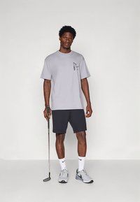 Šviesiai pilka medvilninė trumpomis rankovėmis marškinėliai su juodu grafiniu piešiniu krūtinėje, derinami su tamsiomis šortais ir pilkais sportiniais batais. Rankose laikomas golfo klubas.