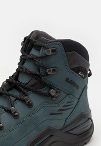 Stivale da trekking verde in pelle con dettagli in rete nera, dotato di occhielli metallici, superficie texturizzata e una spessa suola in gomma nera per una migliore aderenza.