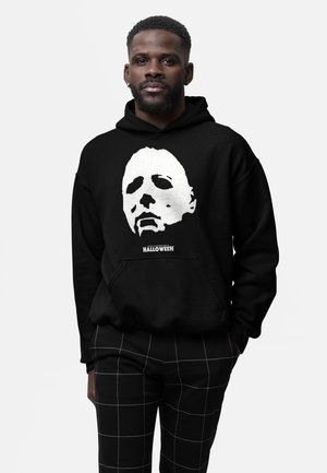 Uomo con felpa nera con cappuccio, stampa della maschera bianca di Michael Myers e testo "John Carpenter’s Halloween", abbinata a pantaloni neri a quadri.