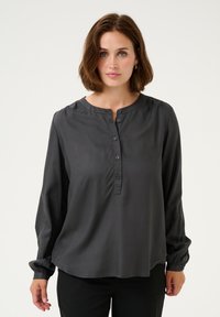 Donkergrijze blouse met knoopsluiting, rond de hals, lange mouwen met verzamelde manchetten en een gladde, zachte textuur. Vier knopen zichtbaar op de borst.