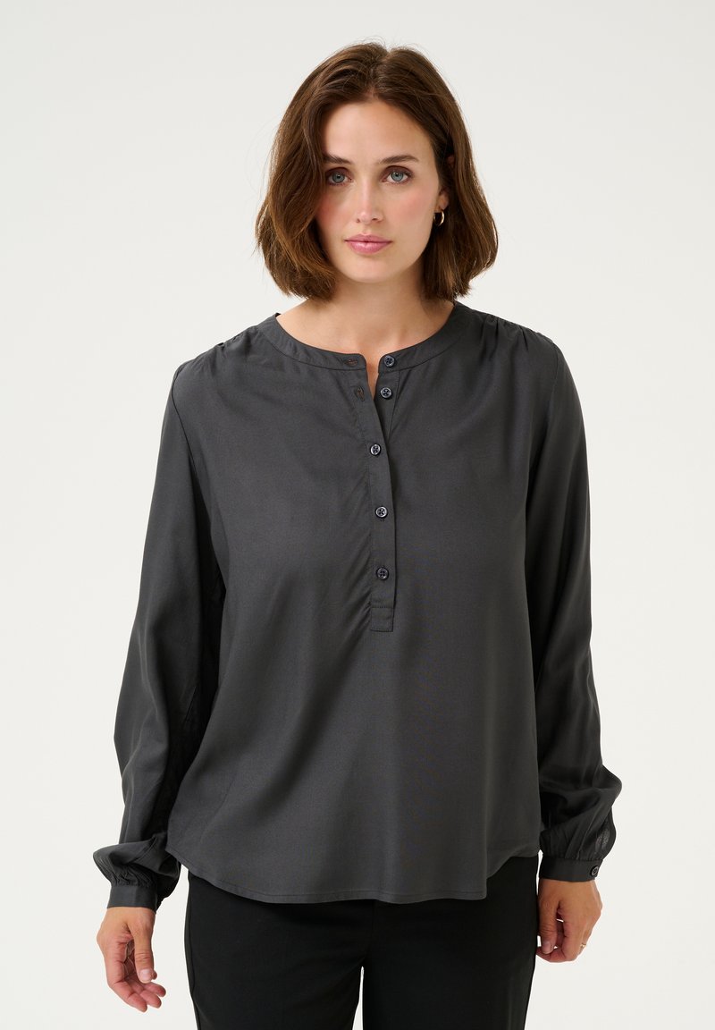 Donkergrijze blouse met knoopsluiting, rond de hals, lange mouwen met verzamelde manchetten en een gladde, zachte textuur. Vier knopen zichtbaar op de borst.