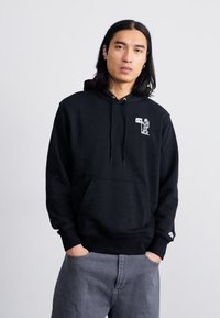 Sweat à capuche en coton noir avec une poche kangourou à l'avant, un capuchon à cordon et un accent graphique sur la poitrine. Associé à un pantalon gris.