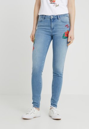 Femme portant un jean skinny bleu clair avec des patchs floraux rouges et des baskets blanches, debout devant un fond uni et clair.