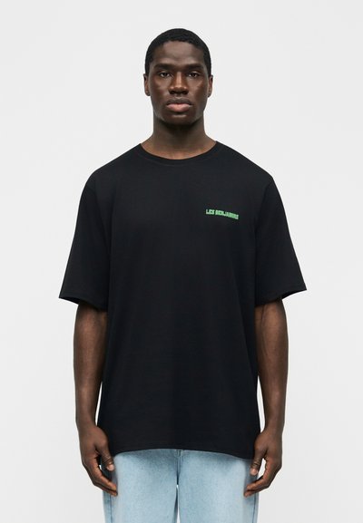 Νεαρός άνδρας με σκουρόχρωμο δέρμα που φοράει μαύρο φαρδύ T-shirt με πράσινο κείμενο "LES BENJAMINS" και ανοιχτόχρωμο μπλε τζιν, στέκεται μπροστά σε απλό φόντο.