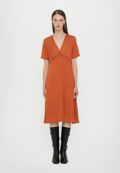 Roodbruin oranje jurk met korte mouwen, V-hals en knoopdetail. Vloeiende silhouet, gestructureerde stof, gecombineerd met kniehoge zwarte laarzen.