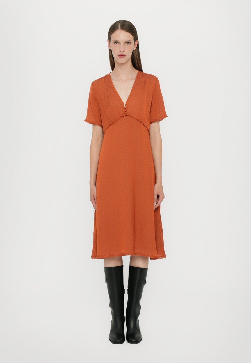 Vestido color óxido con mangas cortas, escote en V y detalle de botones. Silueta fluida, tejido texturizado, combinado con botas negras hasta la rodilla.