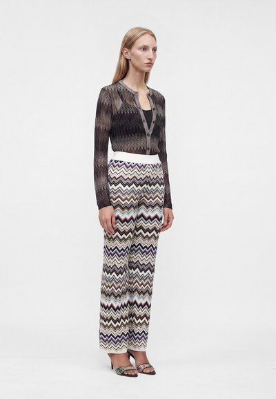 Femme portant un cardigan noir à motifs, un pantalon large à motif chevrons et des talons ouverts, debout devant un fond clair uni.