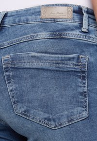 Jeans de denim de color azul claro con un diseño clásico de cinco bolsillos, que presentan una etiqueta rectangular con "Soccer Denim" y detalles de costura en contraste.