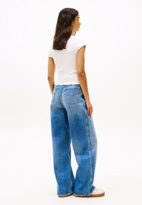 Jeans in denim chiaro a vita alta, ampi in fondo, con una texture sfumata, tasche posteriori e una camicia bianca con orlo scallop che accentua la vita.