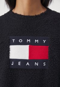 Pull noir texturé avec un patch rectangulaire affichant un motif rouge, blanc et bleu marine et le texte "Tommy Jeans" sur la poitrine.