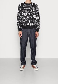 Maglione nero a maglia con simboli grafici bianchi, abbinato a jeans di denim scuro e sneakers con dettagli marroni. Presenta un collo rotondo e polsini a costine.