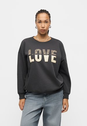 Femme debout, portant un sweat noir avec le mot "LOVE" en grandes lettres dorées et à imprimé animal, et un jean bleu clair, fond neutre.