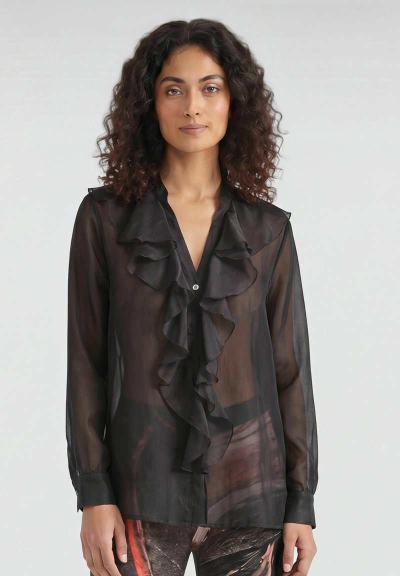 Femme aux cheveux bouclés portant un chemisier noir transparent à manches longues avec des volants sur le devant et un pantalon à motifs.