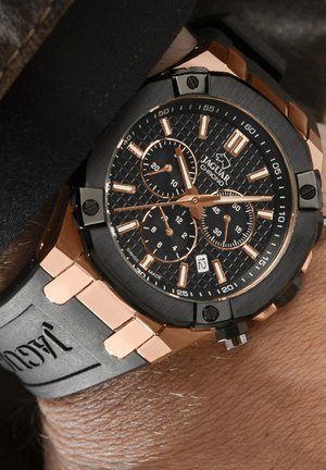 Chronograaf horloge met een zwarte gestructureerde wijzerplaat, roségouden accenten en een zwarte siliconen band met gegraveerde branding.