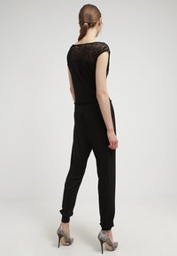 Mono negro con espalda acentuada en encaje, piernas ajustadas, corte relajado y combinado con tacones de serpiente plateados.