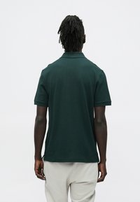 Mężczyzna z dredami stoi tyłem, ubrany w ciemnozieloną koszulkę polo z krótkim rękawem i jasnobeżowe spodnie na jednolitym białym tle.