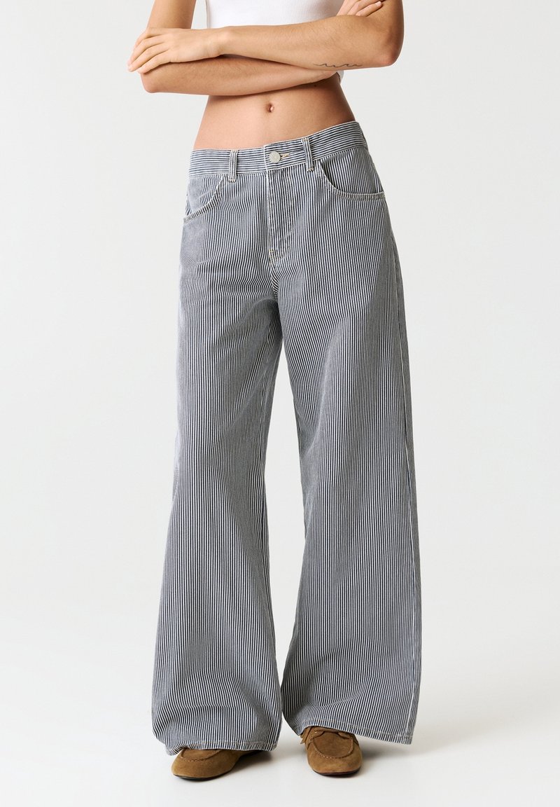Femme portant un pantalon taille haute à jambes larges, rayé noir et blanc, avec des chaussures marron, debout devant un fond clair uni.