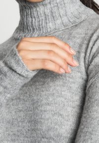 Pull à col roulé gris en maille avec texture côtelée au col et aux poignets, présentant une finition douce et légèrement duveteuse.