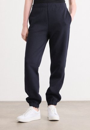 Marineblå joggebukser laget av myk stoff, med elastisk midje, sidelommer og innsnevrede mansjetter, sammen med hvite joggesko.