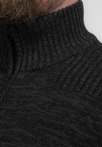 Jersey de cuello alto oscuro y acanalado. La tela presenta un patrón texturizado de punto en varios tonos de negro.