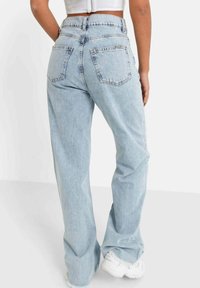 Persona che indossa jeans a vita alta di un blu chiaro e sneakers bianche, vista da dietro su uno sfondo uniforme.