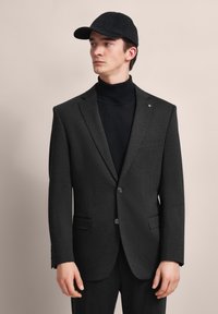 Schwarze, maßgeschneiderte Herrenanzugjacke aus einem weichen Stoff, mit zwei Knöpfen, seitlichen Taschen und einem modernen Schnitt, getragen über einem schwarzen Rollkragenpullover.