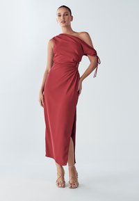 Asimetriska sarkanā kleita no gludas auduma, ar savākto sānu, augstu šķēlumu augšstilbā un pleciem atsegta dizainu. Akcentēta ar plānām siksniņām.