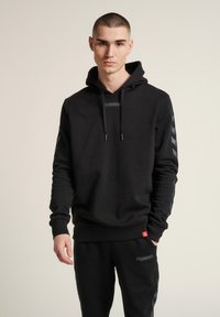 Schwarzer Kapuzenpullover mit glatter Textur, ausgestattet mit einem Logo auf der Vorderseite und gemusterten Akzenten an den Ärmeln; kombiniert mit passenden schwarzen Jogginghosen.