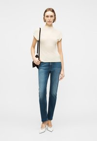 Γυναίκα με κρεμ κοντομάνικο τοπ, μπλε τζιν, λευκά μυτερά παπούτσια και κρατώντας μια μικρή μαύρη τσάντα ώμου, μπροστά σε απλό φόντο.