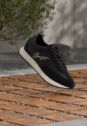 Baskets noires avec semelle blanche et logo « Boss » sur le côté, placées en extérieur sur des dalles en bois avec un feuillage vert en arrière-plan.