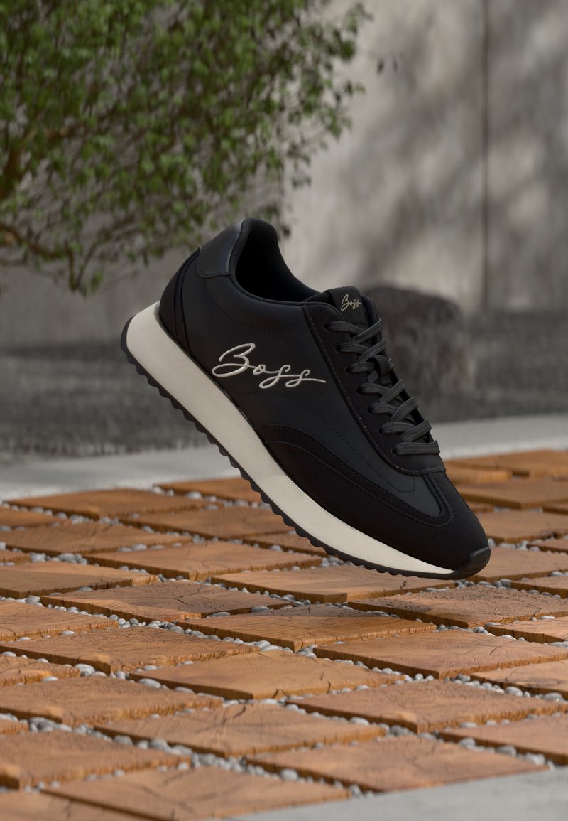 Baskets noires avec semelle blanche et logo « Boss » sur le côté, placées en extérieur sur des dalles en bois avec un feuillage vert en arrière-plan.