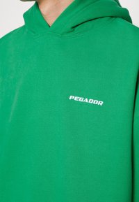 Felpa verde in cotone con tasca a marsupio frontale e logo bianco "PEGADOR" sul petto, realizzata in tessuto testurizzato e dal taglio rilassato.