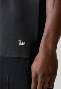 Nahaufnahme eines dunkelhäutigen Arms neben einem schwarzen Shirt mit einem kleinen weißen bestickten Logo am Saum und einer schwarzen Hose mit Tasche.