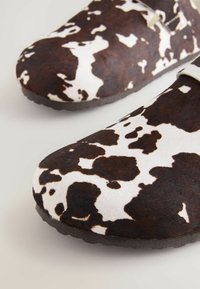 Zuecos con estampado de vaca marrón y blanco, con una superficie suave y texturizada y puntera redondeada. Cuentan con una suela resistente para un buen agarre.