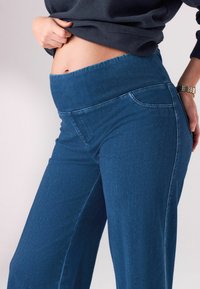Pantaloni din denim albastru cu talie înaltă, prevăzuți cu un brâu elastic lat, având o textură netedă și un design minimalist. Croială ajustată cu buzunare laterale.