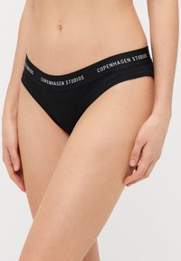 Svart, ribbet bikini-bunn med bred midjebånd med "COPENHAGEN STUDIOS"-logo i hvitt. Glatt tekstur og snug passform.