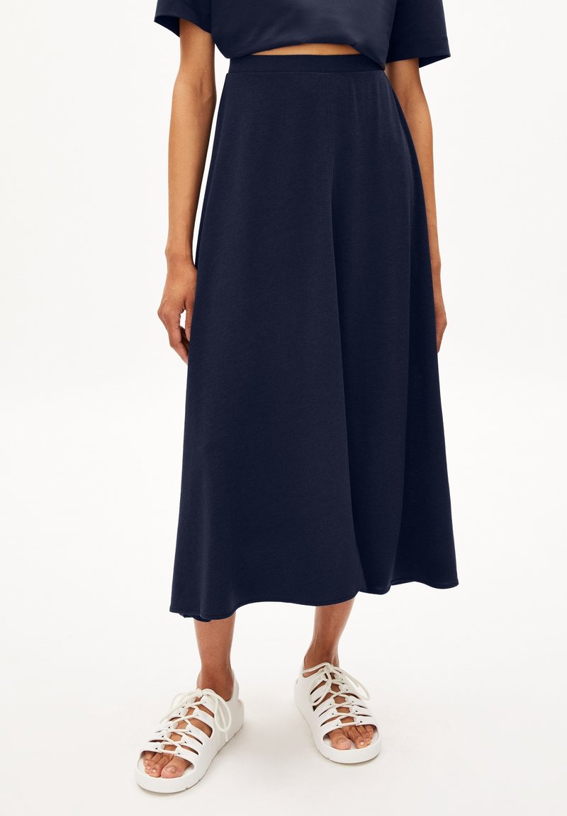 Femme portant une jupe midi bleu marine associée à des sandales blanches à lacets et orteils ouverts, debout devant un fond blanc uni.
